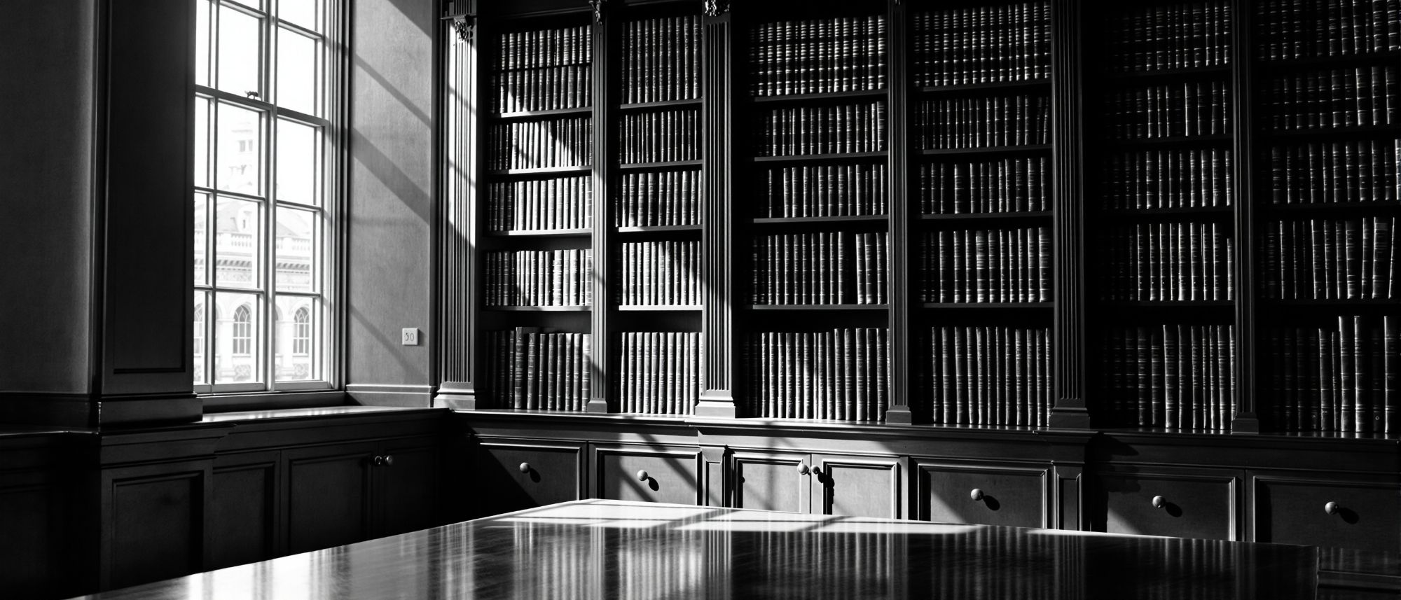 Biblioteca jurídica con estanterías de volúmenes encuadernados y luz lateral
        entrando por un ventanal alto.